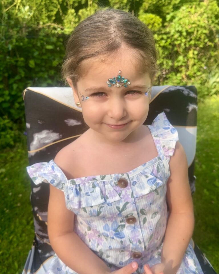 Young girl with jeweled face decoration sits on a patterned chair outdoors, wearing a floral dress, with greenery in the background—ready to shine like meg a star at your next party services event.