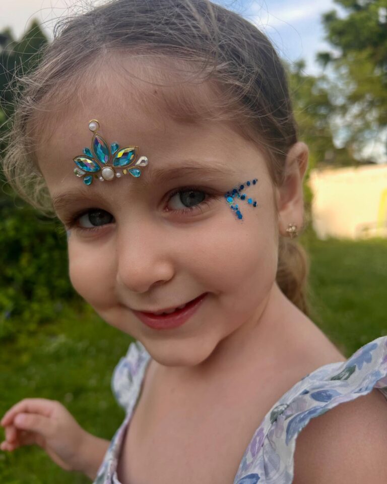 Young girl with face jewels, including a decorative gem on her forehead and blue gems on her cheek, smiles outdoors in a garden setting—perfect for a meg a star party services celebration.