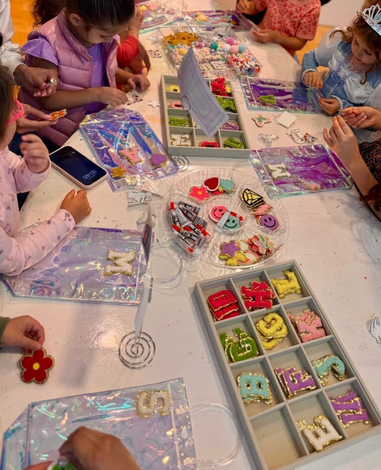 Children sit around a table decorating clear bags with colorful patches and shapes, enjoying a meg a star craft activity. Various supplies and decorated items fill the table, showcasing party services in action.