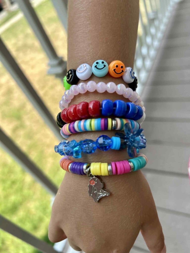 A child's arm wearing multiple colorful beaded bracelets, some with smiley faces and one with a penguin charm, outdoors near a railing.