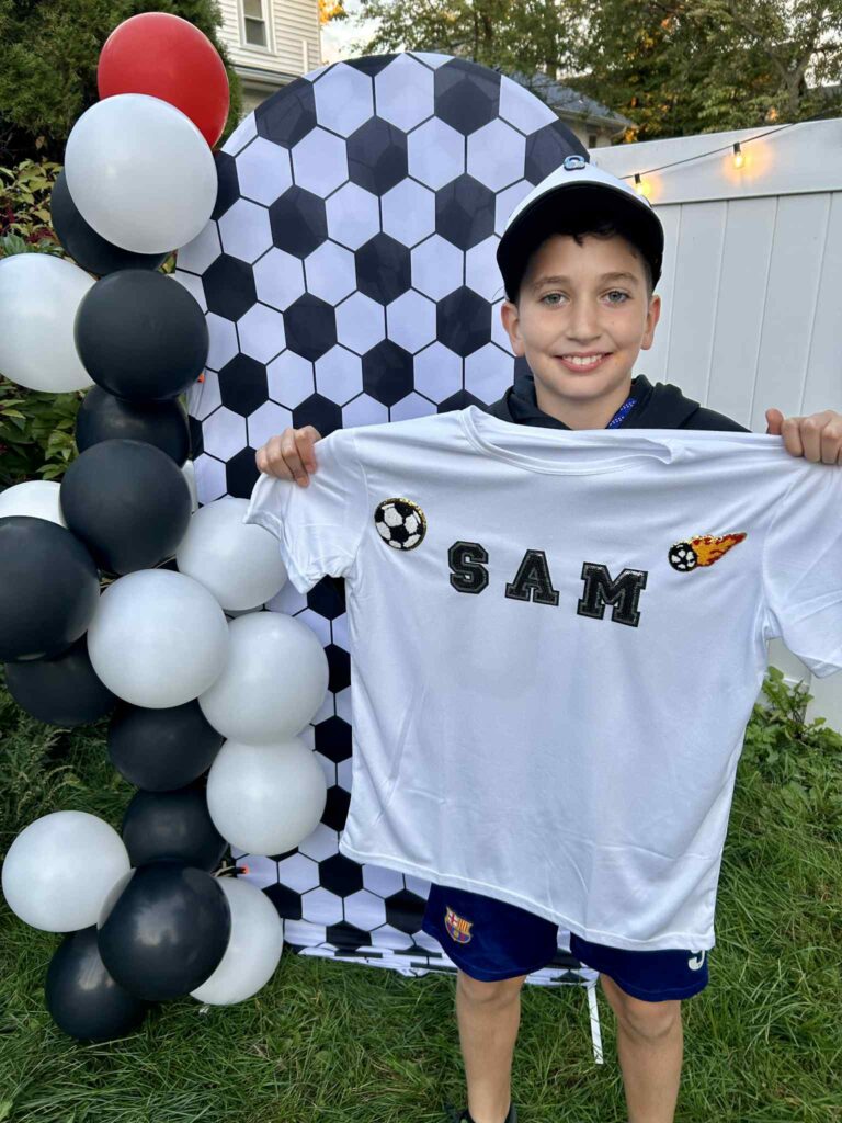 A boy stands outdoors holding a personalized soccer-themed shirt with "SAM" on it, next to a black-and-white balloon display and a soccer ball-patterned backdrop.