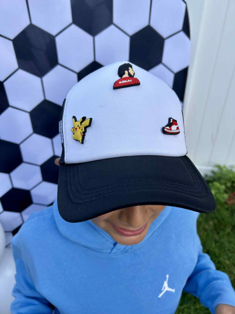 A child wearing a black and white cap with pins of Pikachu, a red shoe, and a person, standing in front of a black-and-white hexagon patterned backdrop.