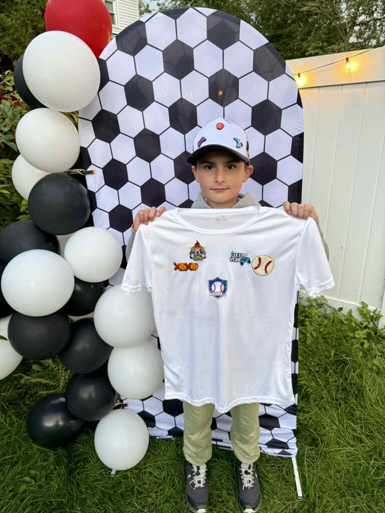 A boy wearing a white cap holds up a white T-shirt with sports-themed patches. He stands in front of a black-and-white soccer ball backdrop and balloon arrangement.