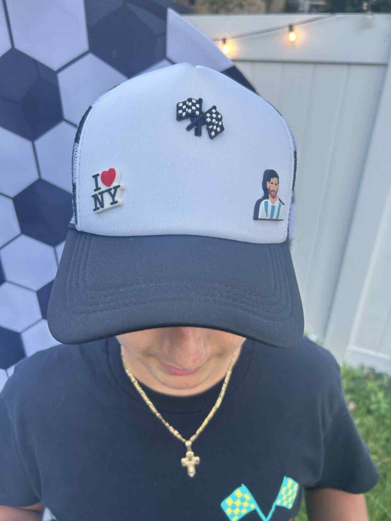 A person wearing a black and white cap decorated with pins: a checkered flag, "I ♥ NY," and a soccer player. The person also wears a gold cross necklace and a black T-shirt.